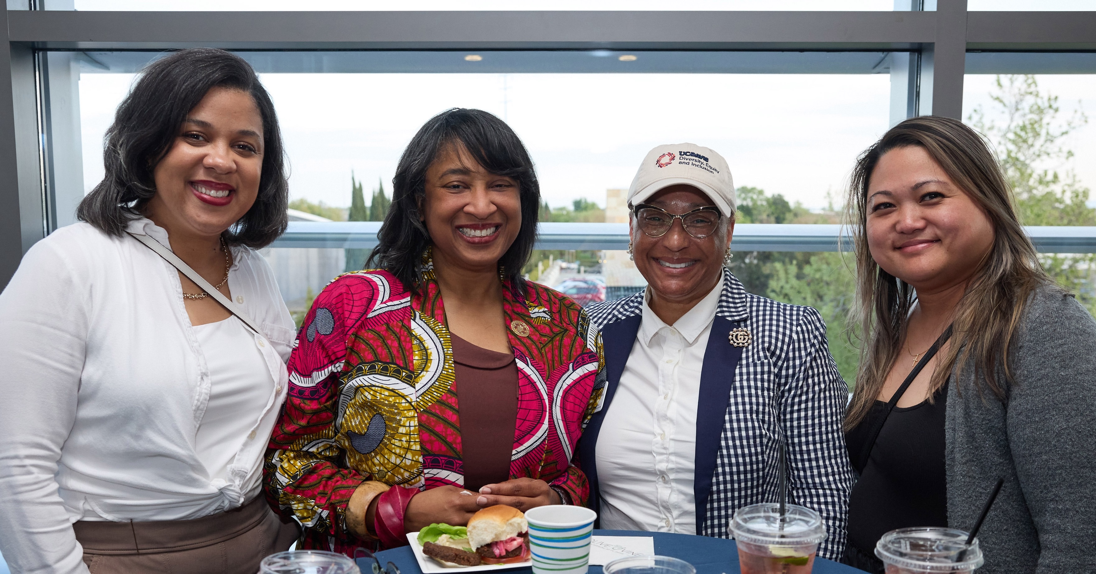 Four women smiling and standing around a table with food and drinks, in front of a large window with a view outside.